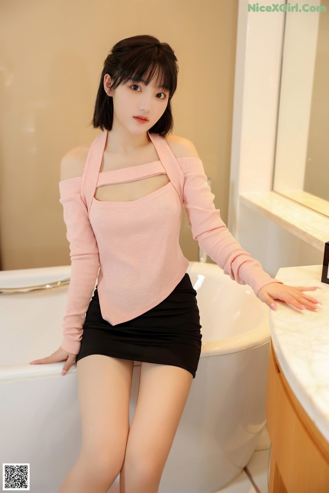 A woman in a pink sweater and black skirt sitting in a bathtub.