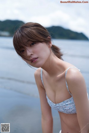 A woman in a white tank top and red shorts crouching on a rock by the ocean.