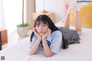 A woman in a blue shirt and gray skirt sitting on a bed.