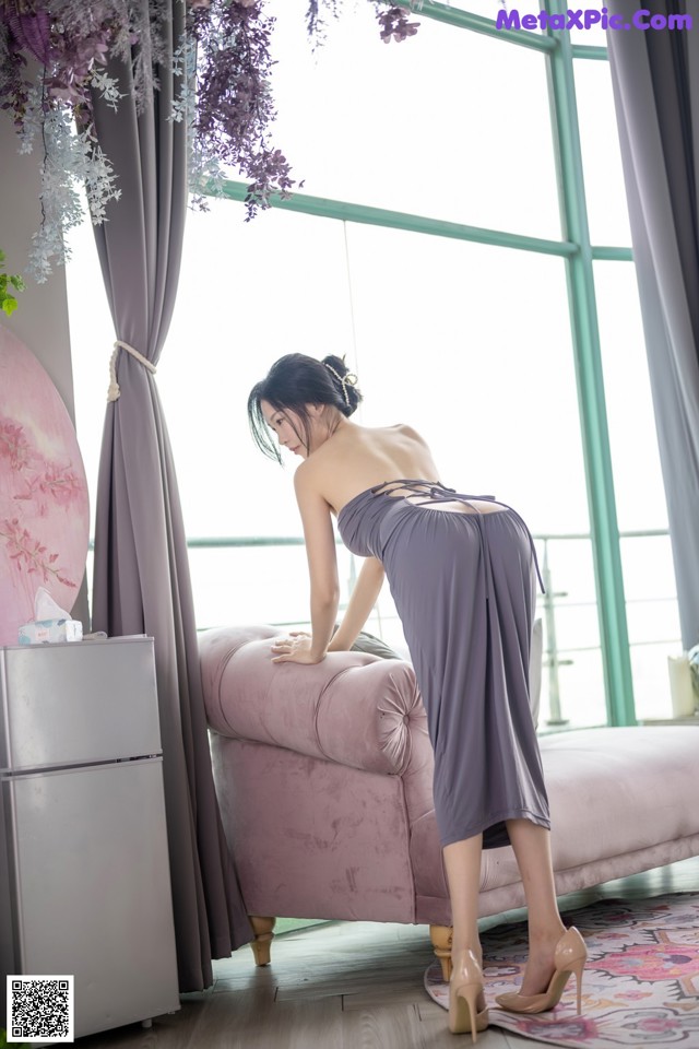 A woman in a gray dress bending over a pink couch.